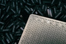 A close-up view of a table covered with small, black, rectangular objects each containing words like 'frame', 'panic', 'junior', and more. Positioned up front is a beige, grid-like chart with columns labeled 1st to 6th and rows marked A to P. A caution message at the top of the chart explains that viewing it gives the viewer full access.