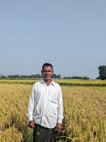 Smiling farmer standing proudly beside lush green crops under a clear blue sky.
