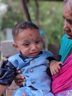 A young child dressed in a light blue outfit with dark blue details is being held by an elderly person wearing a pink and teal garment. The child appears to be crying or upset, with a visibly distressed expression. The scene takes place in a natural, outdoor setting with soft blurred greenery in the background.