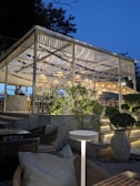 An outdoor seating area with a modern, well-lit pergola during twilight. The structure features hanging lights and international flags as decorations. The seating includes comfortable chairs with cushions around tables. There is lush greenery surrounding the space, adding to the ambiance. A person is visible near the counter area.