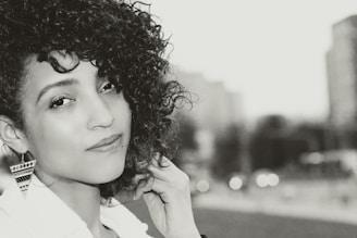 A portrait of a person with curly hair and an enigmatic smile, looking directly at the camera. The focus is on their face, with a blurred background of what appears to be an urban environment. The monochrome filter adds a classic and timeless feel to the image. The person is wearing large, patterned earrings and a light-colored top.