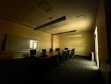 A dimly lit conference room with a long table surrounded by several office chairs. A large whiteboard is mounted on the wall, and light filters through a window, casting shadows on the carpeted floor.