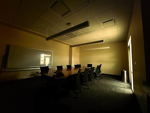 A dimly lit conference room with a long table surrounded by several office chairs. A large whiteboard is mounted on the wall, and light filters through a window, casting shadows on the carpeted floor.
