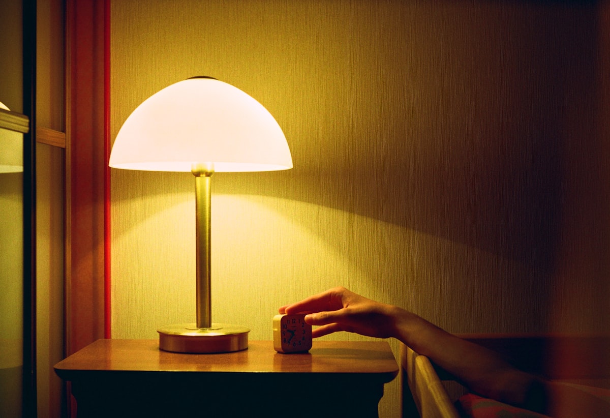 A person's hand reaching for a clock on a nightstand by lamplight, the nighttime cutoff moment