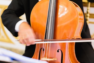 A person is playing a cello, with the musician's hand in motion holding the bow across the strings. The cello has a polished, wooden surface, and the player's attire is formal, suggesting a classical music performance setting.