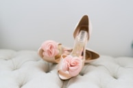 A pair of elegant high-heeled shoes with decorative rose-like embellishments at the toes, placed on a soft, tufted surface.