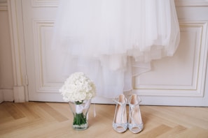 A white wedding dress is elegantly hung on a white paneled door. Below the dress, a clear glass vase holds a bouquet of white roses. Next to the vase, a pair of sparkling high-heeled shoes with intricate details is placed on the wooden floor.