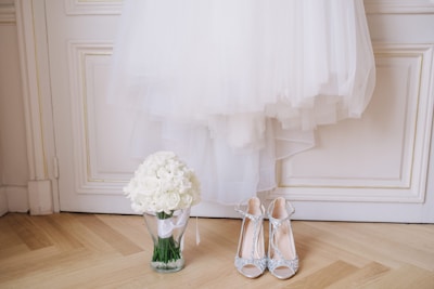 A white wedding dress is elegantly hung on a white paneled door. Below the dress, a clear glass vase holds a bouquet of white roses. Next to the vase, a pair of sparkling high-heeled shoes with intricate details is placed on the wooden floor.