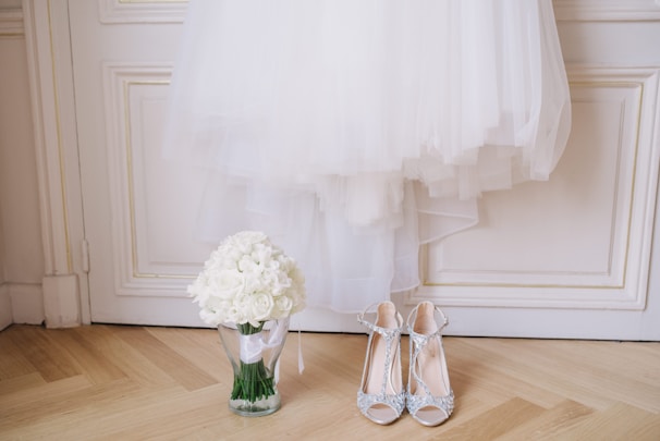 A white wedding dress is elegantly hung on a white paneled door. Below the dress, a clear glass vase holds a bouquet of white roses. Next to the vase, a pair of sparkling high-heeled shoes with intricate details is placed on the wooden floor.