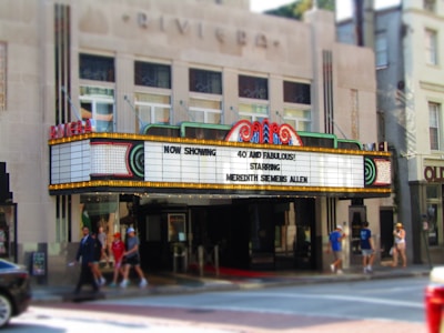 A vintage theater marquee displaying the message 'Now Showing: 40 and Fabulous! Starring Meredith Semmes Allen' in bright lights. The theater exterior features an art deco architectural style with decorative neon accents. People are walking on the sidewalk in front of the theater, and a partial view of a car can be seen on the street.