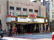 A vintage theater marquee displaying the message 'Now Showing: 40 and Fabulous! Starring Meredith Semmes Allen' in bright lights. The theater exterior features an art deco architectural style with decorative neon accents. People are walking on the sidewalk in front of the theater, and a partial view of a car can be seen on the street.