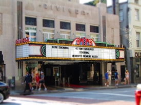A vintage theater marquee displaying the message 'Now Showing: 40 and Fabulous! Starring Meredith Semmes Allen' in bright lights. The theater exterior features an art deco architectural style with decorative neon accents. People are walking on the sidewalk in front of the theater, and a partial view of a car can be seen on the street.