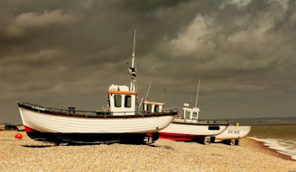Three fishing boats are resting on a pebble beach under a cloudy sky. The boats are painted white with red and black accents and appear to be positioned for maintenance or storage. The shoreline and waves are visible in the background, contributing to a tranquil yet slightly somber atmosphere.