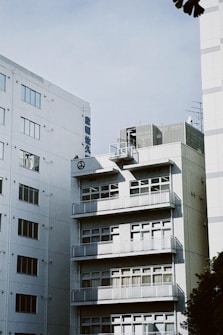 A large, multi-story office building in an urban setting. The structure is comprised of multiple floors with rows of windows and balconies. The building is made of concrete and glass and features a logo and some text in an Asian language on the facade. The sky above is clear with a pale blue tone, and there is a hint of foliage in the lower right corner.