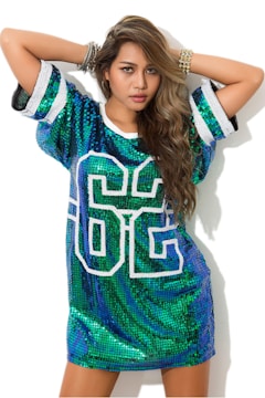 A person is wearing a sequined dress with the number 86 on the front. The dress is predominantly green and blue with sparkly sequins, and the person has long, wavy hair. They have an arm raised above their head and are wearing multiple bracelets.
