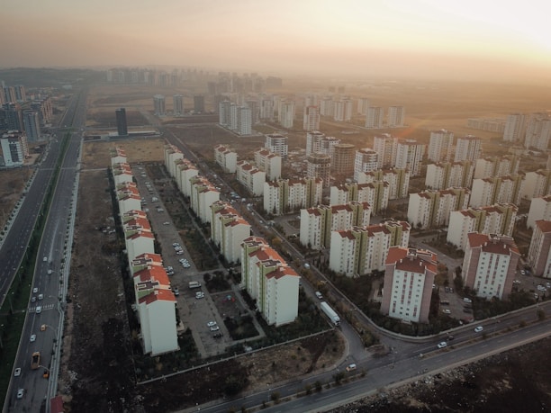Aerial view of a sprawling residential area featuring numerous apartment buildings with red roofs. The buildings are aligned in rows, with some space for roads and paths visible between them. Surrounding the apartment complex is a vast landscape with sparse vegetation and an expansive road running parallel to the buildings. The lighting suggests a sunset, casting a warm glow over the scene.