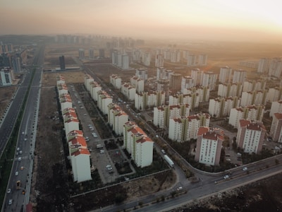 Aerial view of a sprawling residential area featuring numerous apartment buildings with red roofs. The buildings are aligned in rows, with some space for roads and paths visible between them. Surrounding the apartment complex is a vast landscape with sparse vegetation and an expansive road running parallel to the buildings. The lighting suggests a sunset, casting a warm glow over the scene.
