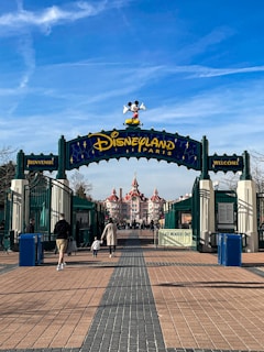 The entrance to Disneyland Paris features an archway with Mickey Mouse atop, flanked by signs in both French and English that say 'Welcome.' In the background, an ornate building with red roofs can be seen, and several people, including a parent and child, walk toward the entrance.