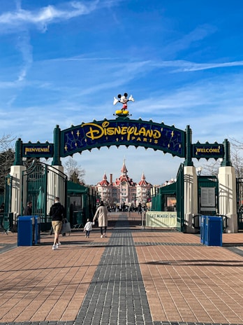 The entrance to Disneyland Paris features an archway with Mickey Mouse atop, flanked by signs in both French and English that say 'Welcome.' In the background, an ornate building with red roofs can be seen, and several people, including a parent and child, walk toward the entrance.
