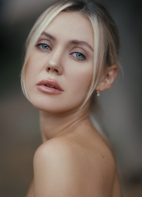 A close-up portrait of a woman with light blonde hair and blue eyes. She is gazing directly at the camera with a neutral expression, her skin is smooth and light in tone. The background is softly blurred, highlighting her features.
