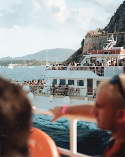 A large boat filled with people enjoying a scenic ride on the water. The boat has multiple decks with passengers taking in the views, socializing, and relaxing. The backdrop includes mountains, a rocky cliffside, other boats, and a blue sky with light clouds. The water appears calm, contributing to the leisurely atmosphere.