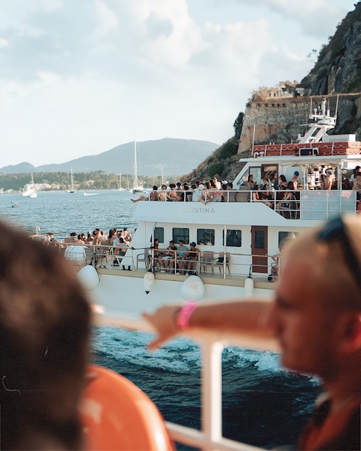 A large boat filled with people enjoying a scenic ride on the water. The boat has multiple decks with passengers taking in the views, socializing, and relaxing. The backdrop includes mountains, a rocky cliffside, other boats, and a blue sky with light clouds. The water appears calm, contributing to the leisurely atmosphere.