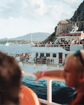 A large boat filled with people enjoying a scenic ride on the water. The boat has multiple decks with passengers taking in the views, socializing, and relaxing. The backdrop includes mountains, a rocky cliffside, other boats, and a blue sky with light clouds. The water appears calm, contributing to the leisurely atmosphere.