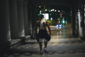 A muscular man is walking through a dimly lit pathway at night. He is wearing a white tank top, black shorts, and white sneakers, with a jacket tied around his waist. The pathway is bordered by columns and leads out to a street with blurred car lights in the background.