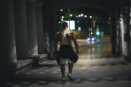 A muscular man is walking through a dimly lit pathway at night. He is wearing a white tank top, black shorts, and white sneakers, with a jacket tied around his waist. The pathway is bordered by columns and leads out to a street with blurred car lights in the background.