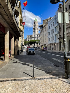 A city street lined with tall, stone buildings on either side. Flags are flying from one of the buildings, and in the distance, there is a grand, historic-looking tower with a green dome. The sky is blue with a few clouds, and the street is relatively empty, with a few parked cars.