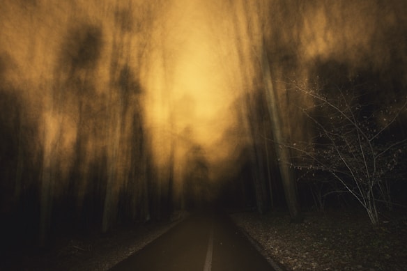 A dark, eerie forest scene with tall trees blurred by moving fog or mist. The scene is illuminated by a yellowish light, casting an ominous glow. A narrow path or road stretches into the distance, surrounded by dense trees on either side. The overall atmosphere conveys mystery and perhaps a touch of the supernatural.