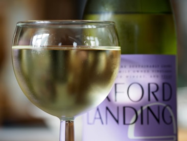 A glass of white wine is filled with a chilled beverage, set against a backdrop featuring a partially visible wine bottle with a purple label. The focus is on the clarity and condensation of the glass.