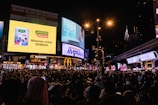 A bustling city scene at night with a large crowd gathered under numerous vibrant advertising billboards. The advertisements feature products and messages in various languages, and streetlights illuminate the area. Shopping and fast-food outlets, such as McDonald's, are visible. The iconic Petronas Twin Towers can be seen in the background.