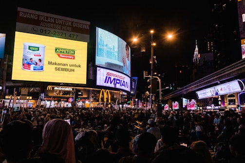 A bustling city scene at night with a large crowd gathered under numerous vibrant advertising billboards. The advertisements feature products and messages in various languages, and streetlights illuminate the area. Shopping and fast-food outlets, such as McDonald's, are visible. The iconic Petronas Twin Towers can be seen in the background.