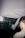 A person in glasses is seated inside a car, with the interior and the rearview mirror visible. The person is looking down at an object or screen. The background through the windshield shows a blurred view of a gas station or convenience store.