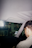A person in glasses is seated inside a car, with the interior and the rearview mirror visible. The person is looking down at an object or screen. The background through the windshield shows a blurred view of a gas station or convenience store.