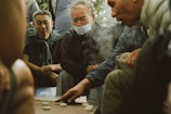 A group of men are gathered around a game board, deeply focused on the game. One man is pointing at the pieces on the board, suggesting an intense moment of strategy. The atmosphere is casual, with the men dressed in everyday clothing, one wearing a mask. The scene appears to be set outdoors, with soft lighting and smoke drifting in the air.