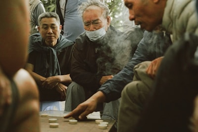 A group of men are gathered around a game board, deeply focused on the game. One man is pointing at the pieces on the board, suggesting an intense moment of strategy. The atmosphere is casual, with the men dressed in everyday clothing, one wearing a mask. The scene appears to be set outdoors, with soft lighting and smoke drifting in the air.