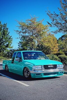 A turquoise Isuzu pickup truck with a lowered suspension is parked in a lot. The vehicle has custom wheels and is situated under a clear blue sky with surrounding green trees and faint autumn colors in the foliage.