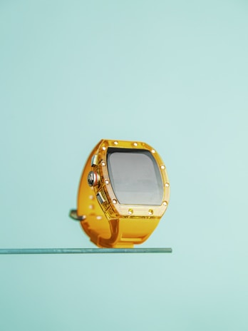 A yellow watch with a large, rectangular face is displayed against a light blue background. The watch is adorned with metallic details, giving it a sleek, modern appearance.