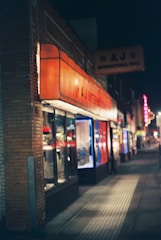 A nighttime street view featuring a series of small shops. The near shop has a bright orange awning with the text 'A J International Mall' on a sign above the store. The shops are illuminated from within, and the street is dimly lit with a few lights visible further in the distance.