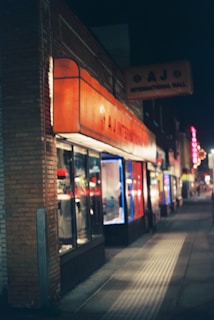 A nighttime street view featuring a series of small shops. The near shop has a bright orange awning with the text 'A J International Mall' on a sign above the store. The shops are illuminated from within, and the street is dimly lit with a few lights visible further in the distance.