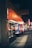 A nighttime street view featuring a series of small shops. The near shop has a bright orange awning with the text 'A J International Mall' on a sign above the store. The shops are illuminated from within, and the street is dimly lit with a few lights visible further in the distance.