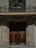 A grand wooden double door with intricate carvings and a brass knocker, set within a stone building facade. Above the door, there is a small balcony with a decorative railing and a window flanked by open wooden shutters. Potted plants are placed on the balcony, adding a touch of greenery.