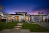A modern, single-story house with clean lines and a contemporary design. The facade features large windows and a wooden double door entry. The house is accented by stone details and illuminated by exterior lighting. The sky is a deep, twilight blue with hints of purple. A well-manicured lawn and a 'For Sale' sign are visible in the foreground.