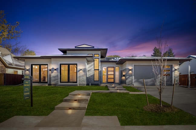 A modern, single-story house with clean lines and a contemporary design. The facade features large windows and a wooden double door entry. The house is accented by stone details and illuminated by exterior lighting. The sky is a deep, twilight blue with hints of purple. A well-manicured lawn and a 'For Sale' sign are visible in the foreground.