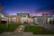 A modern, single-story house with clean lines and a contemporary design. The facade features large windows and a wooden double door entry. The house is accented by stone details and illuminated by exterior lighting. The sky is a deep, twilight blue with hints of purple. A well-manicured lawn and a 'For Sale' sign are visible in the foreground.