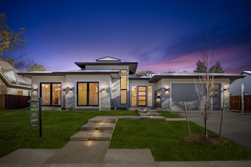 A modern, single-story house with clean lines and a contemporary design. The facade features large windows and a wooden double door entry. The house is accented by stone details and illuminated by exterior lighting. The sky is a deep, twilight blue with hints of purple. A well-manicured lawn and a 'For Sale' sign are visible in the foreground.