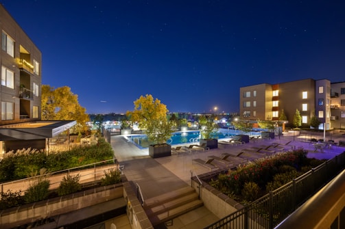 A modern apartment complex at night with a swimming pool area illuminated by artificial lights. The scene includes lounge chairs, potted plants, and well-maintained landscaping. The sky is clear and dark, typical of an evening setting.