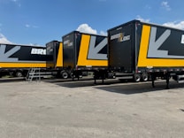 Several large black and yellow logistics trailers are parked in a lot under a clear blue sky. The trailers have bold patterns and the words 'Bros Log' displayed prominently on their sides. A small staircase is next to one of the trailers.